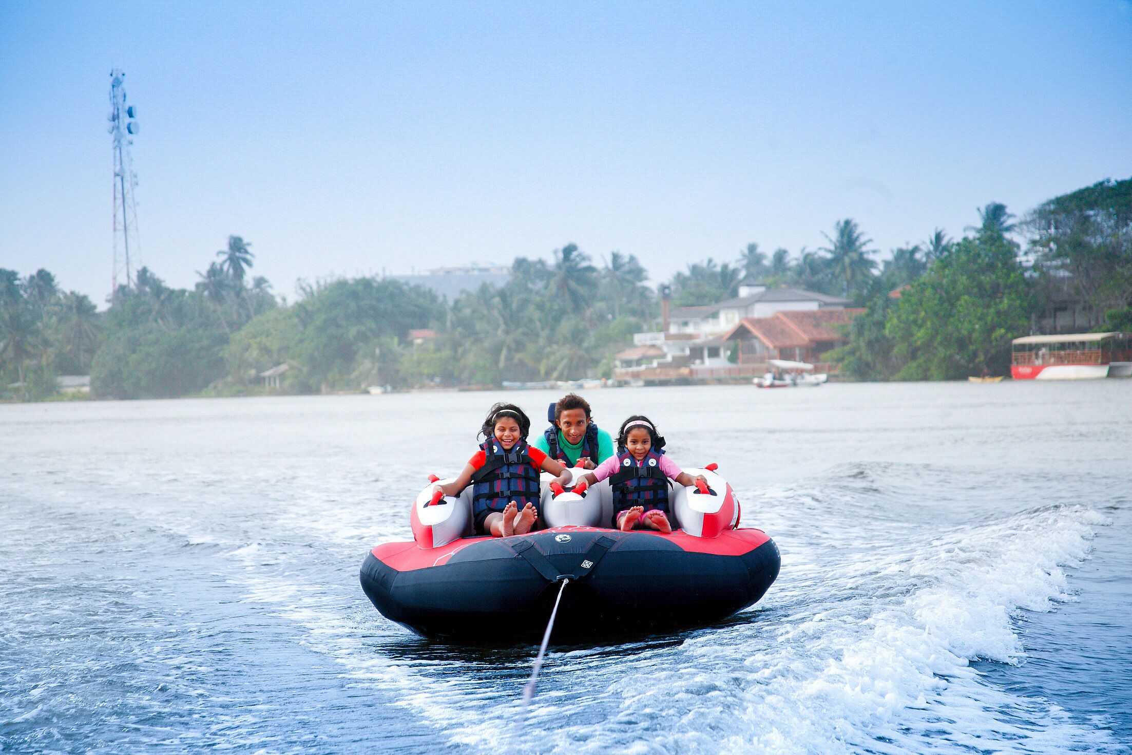 A watercraft with three passengers, a clear sky, and a distant shoreline with buildings and palm trees.