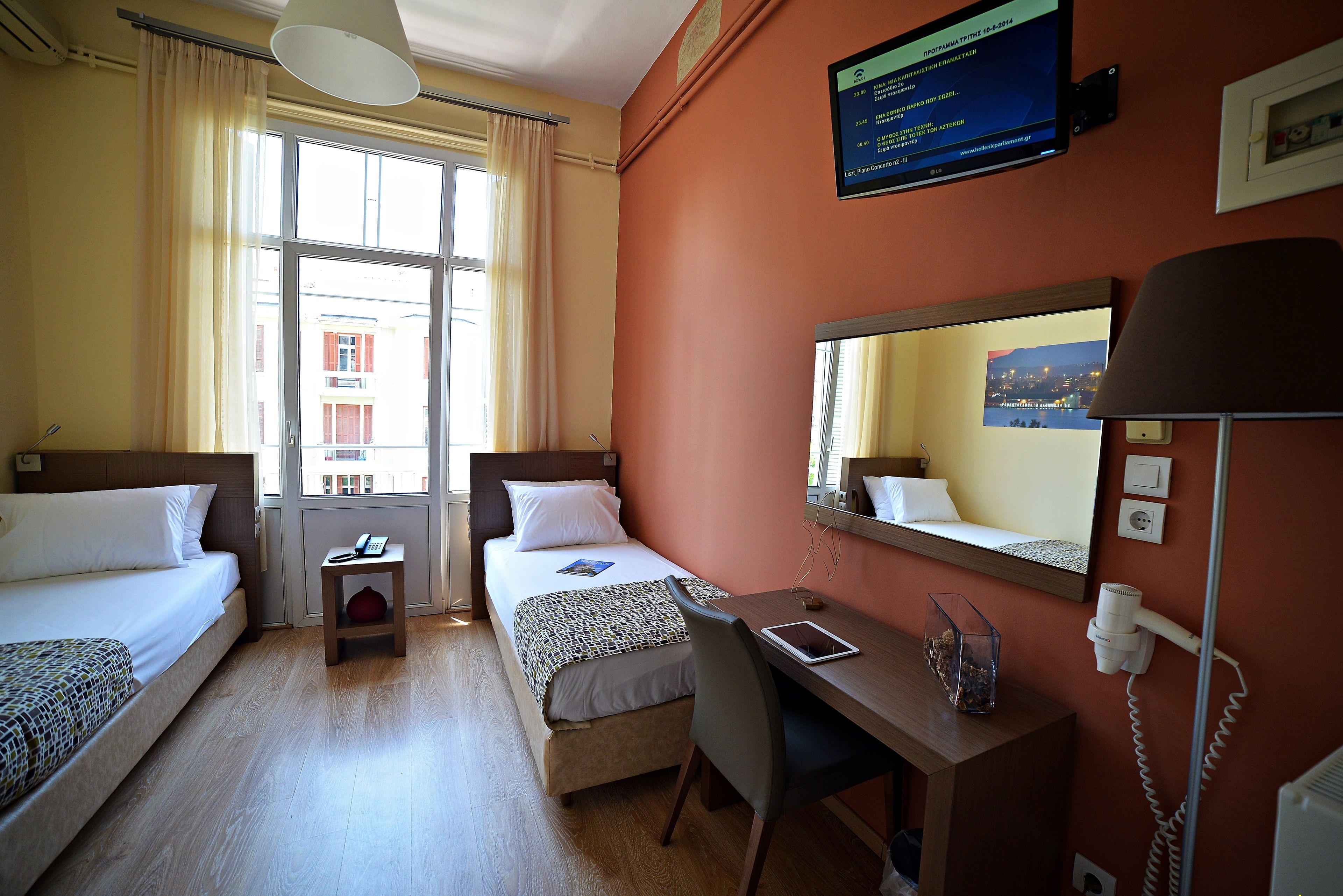 A hotel room with two beds, a desk, a chair, a lamp, a mirror, and a window with curtains.