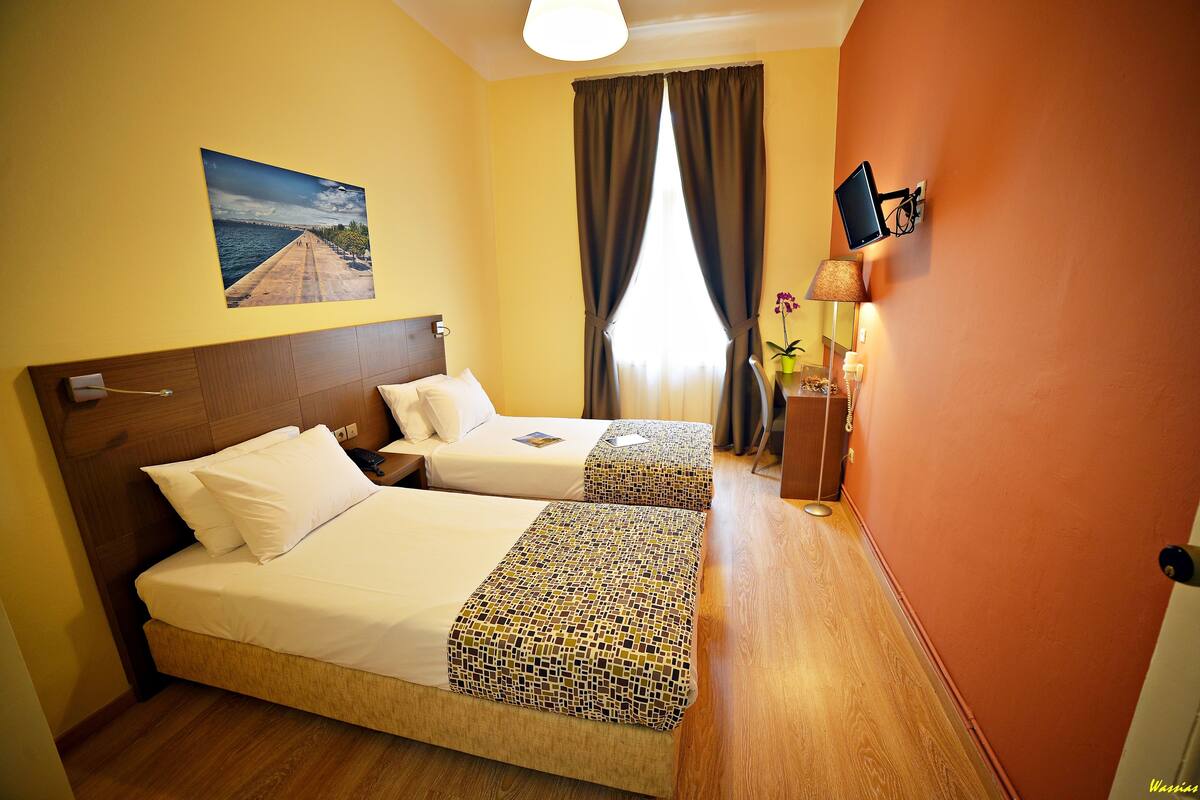 Une chambre d’hôtel avec deux lits, une grande fenêtre avec des rideaux, un téléviseur fixé au mur et un tableau représentant une jetée.