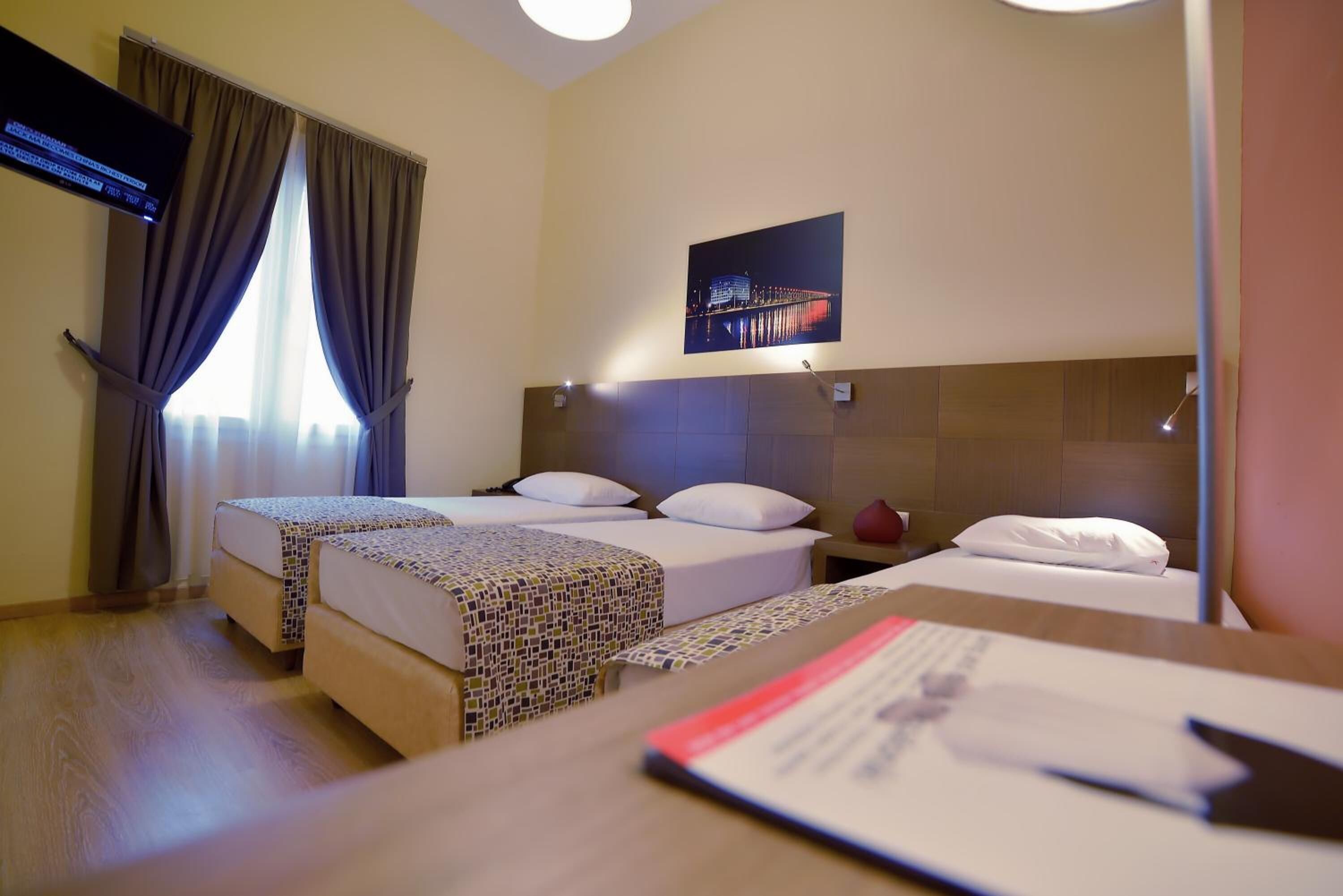 A hotel room with two beds, a flat-screen TV, a painting of a cityscape, and a wooden desk with a red object on it.