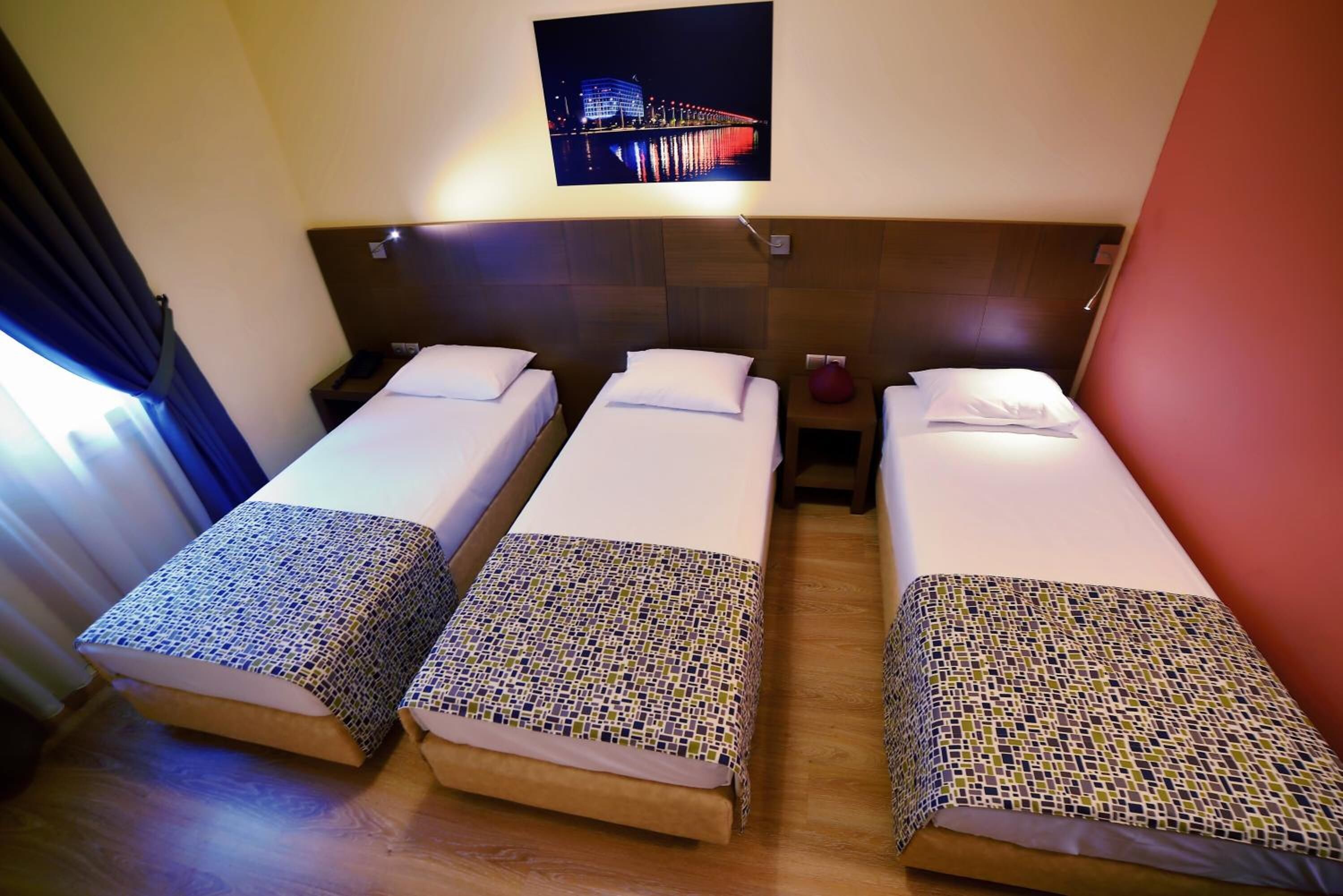 Una camera d'albergo con tre letti singoli, un comodino con una lampada e una TV a muro che mostra una città illuminata di notte.