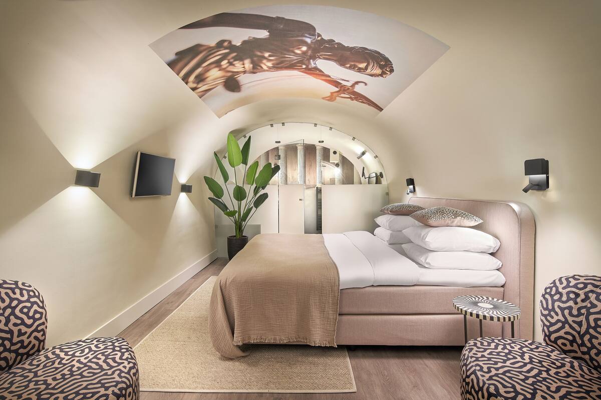 A modern bedroom with a curved ceiling, a bed with a beige blanket, patterned chairs, a small side table, and a potted plant.