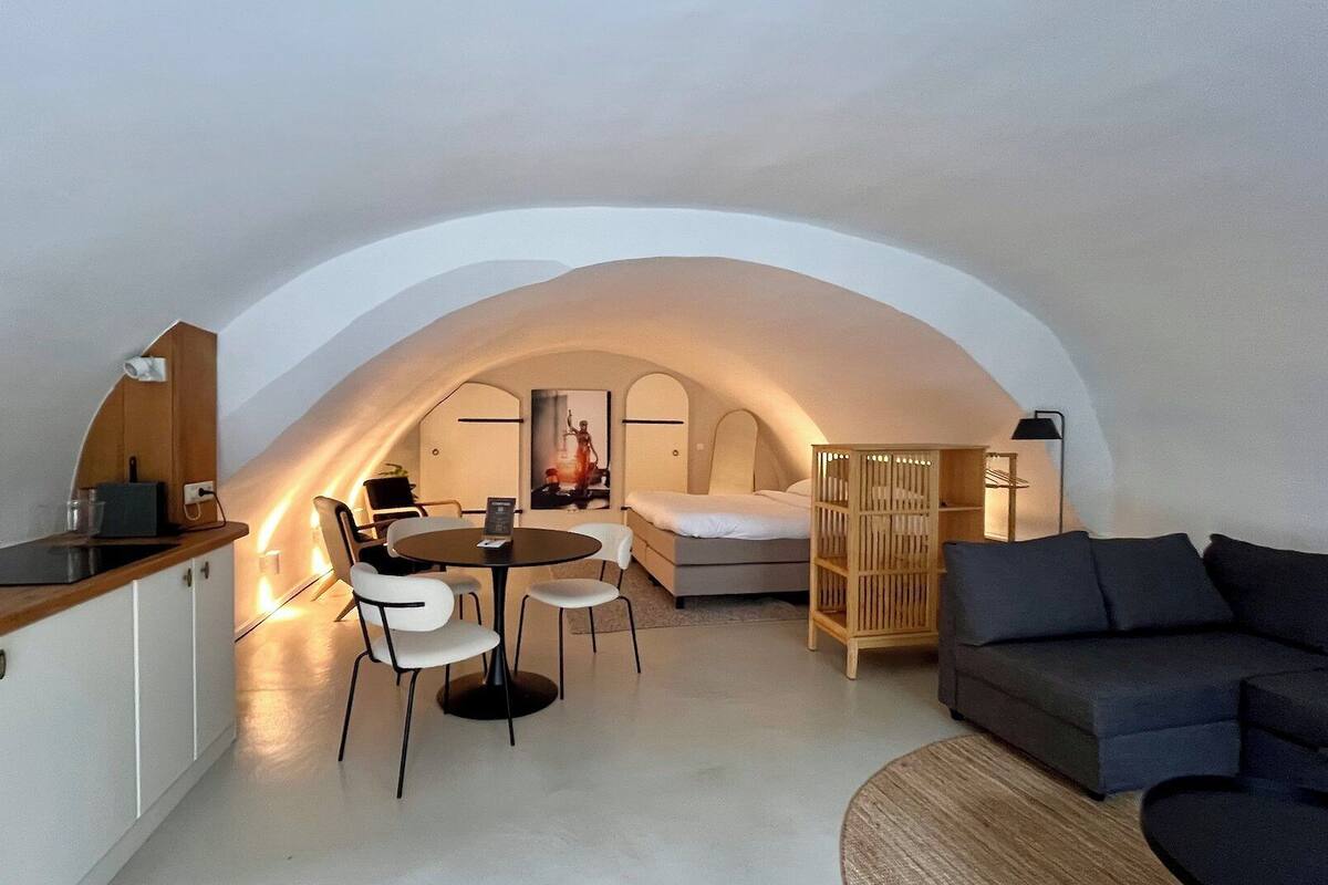 A compact, modern loft bedroom with a curved ceiling, a small dining area, a sofa, and a wooden cabinet.