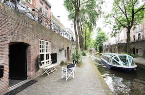 A narrow canal with a boat docked on the right, a brick building on the left with a door and windows, and a bench in the foreground.