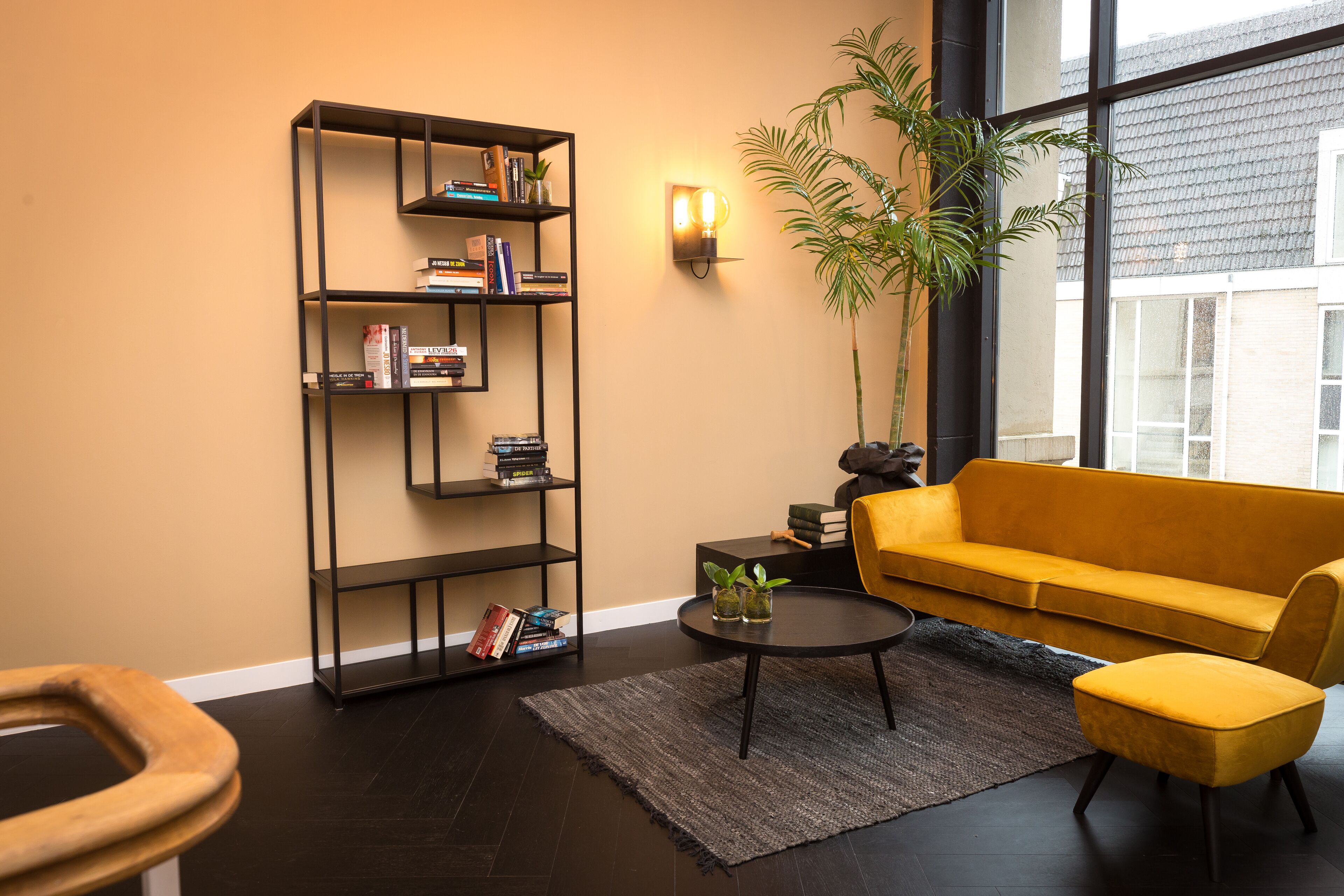 A modern living room with a yellow sofa, a black bookshelf, a potted plant, and a large window.