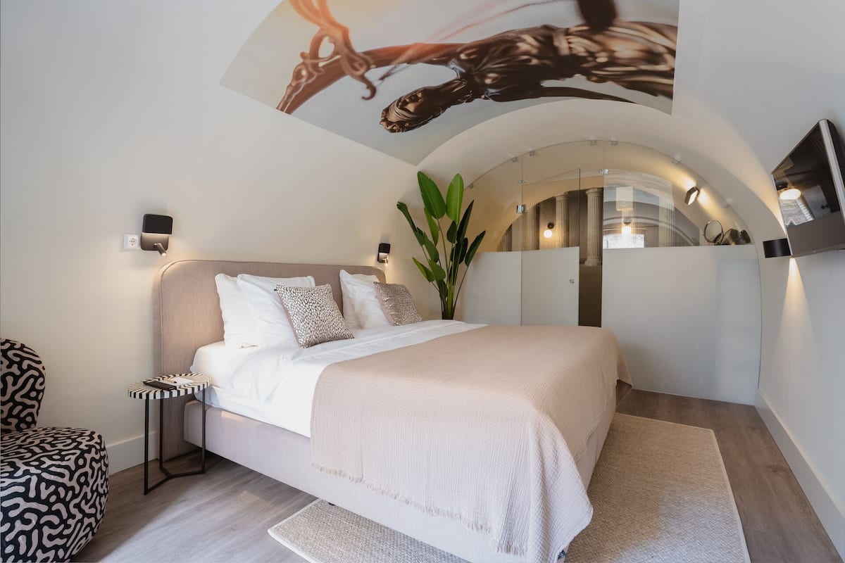 A modern bedroom with a large bed, a side table, a mirror, and a plant.
