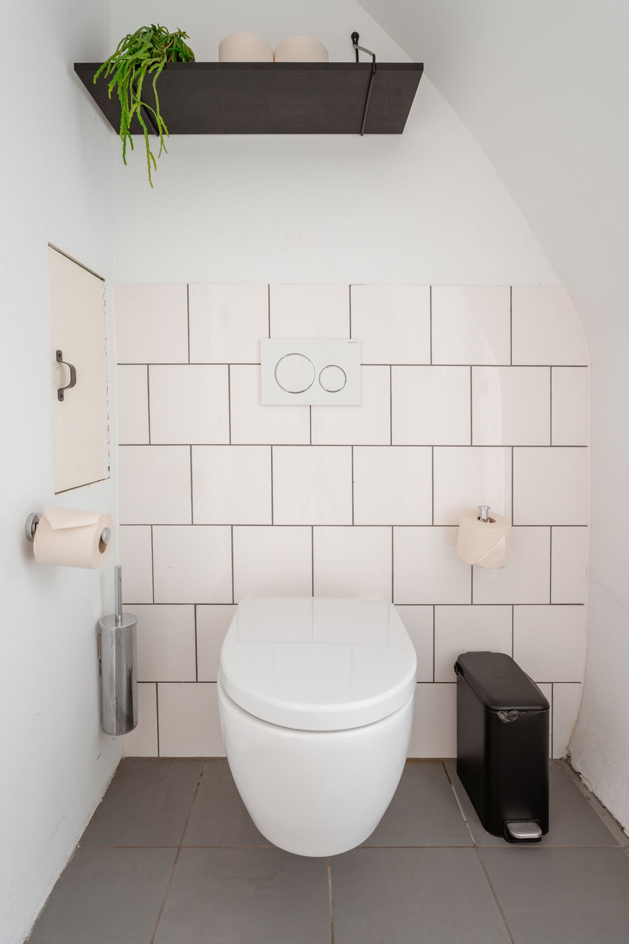 Ein modernes Badezimmer mit Toilette, schwarzem Mülleimer, Toilettenpapierhalter und einer Pflanze auf einem dunklen Regal.
