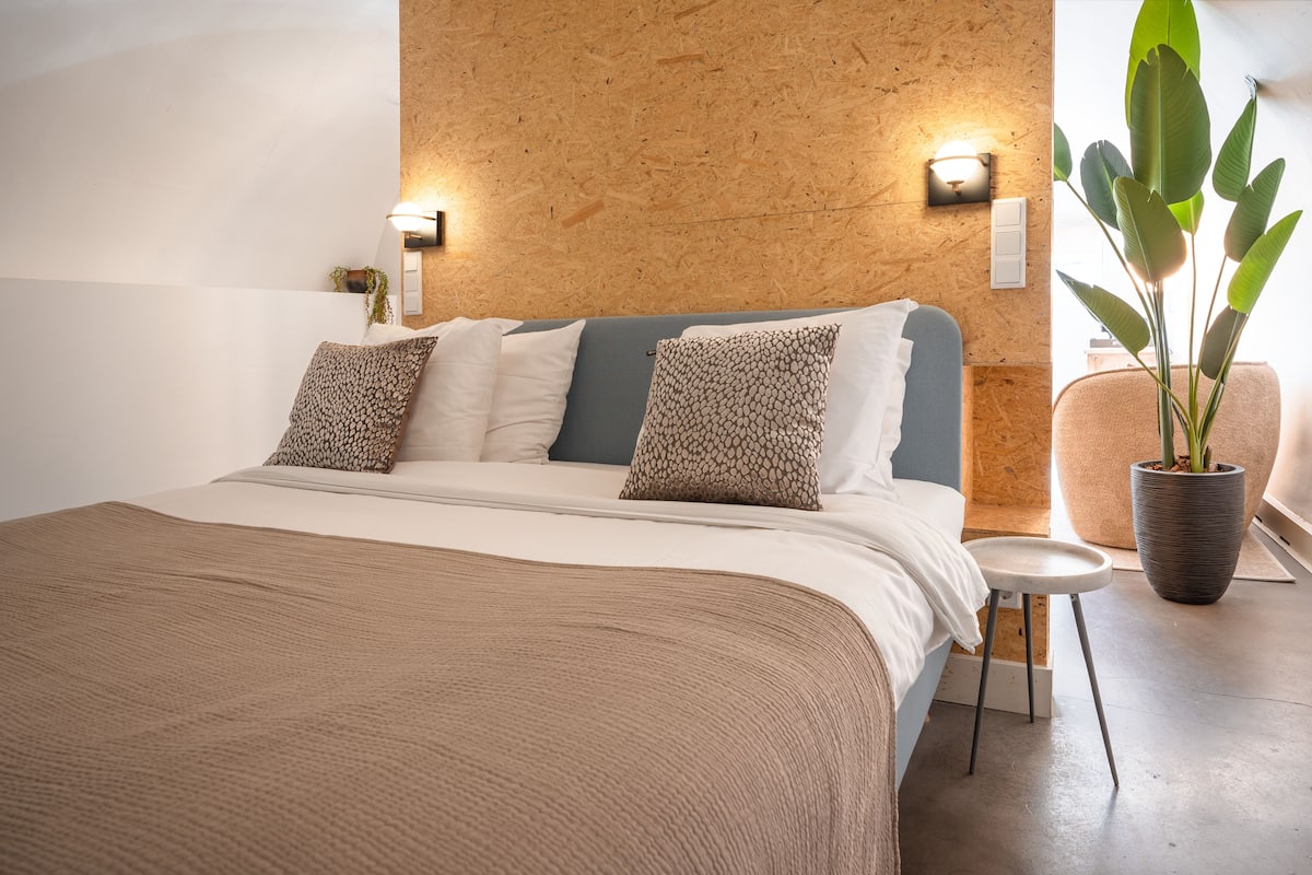 A modern bedroom with a wooden accent wall, a bed with patterned pillows, a small stool, and a large potted plant.
