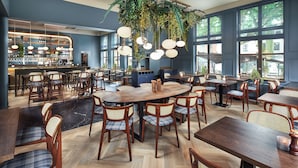A spacious restaurant with wooden tables and chairs, a bar area, and large windows allowing natural light.