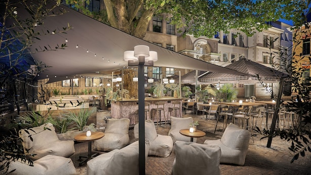 An outdoor seating area with cushioned chairs and tables, surrounded by plants and a large umbrella providing shade.
