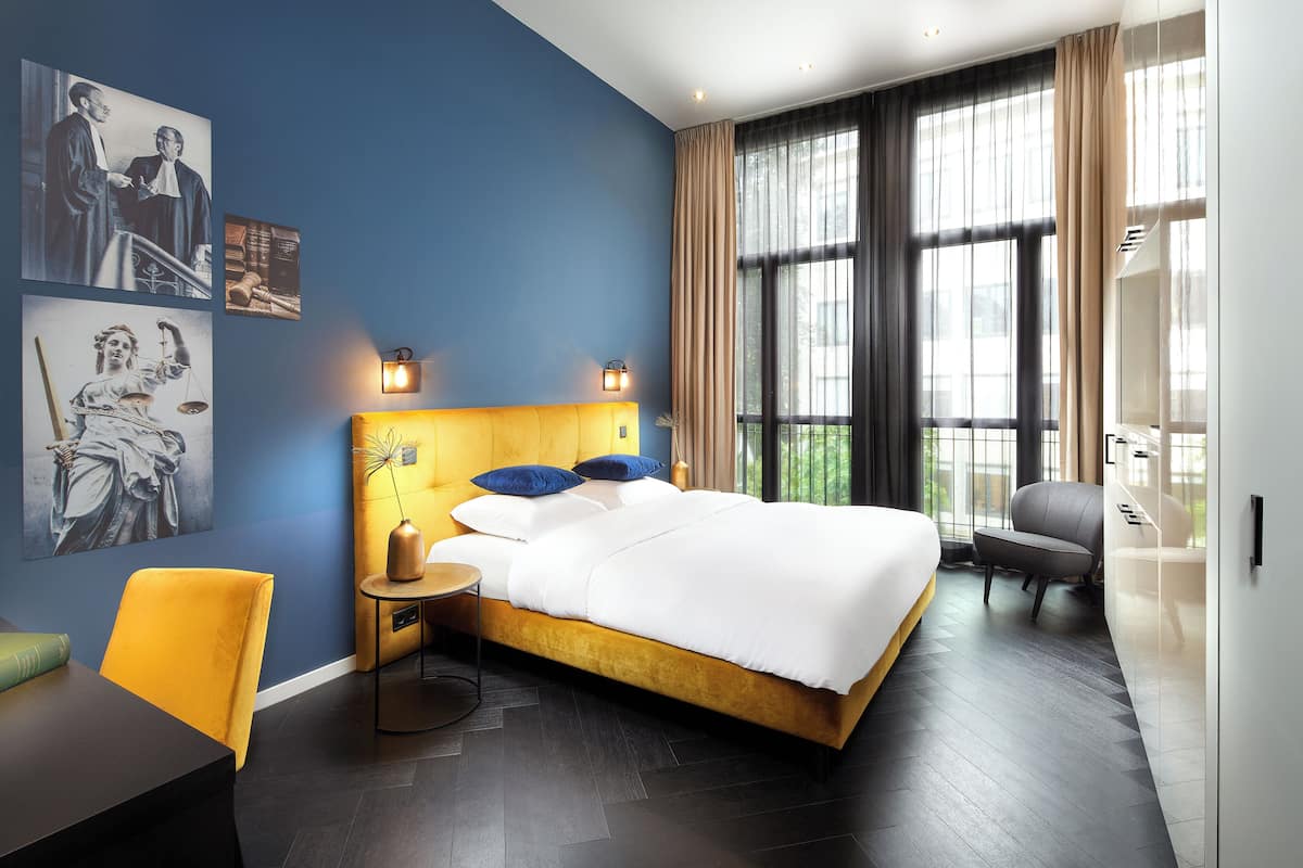 A modern hotel room with a large bed, a desk, and a chair. There are artwork on the walls and a large window with curtains.