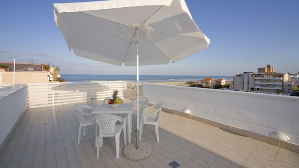 Une terrasse avec un parasol blanc, des chaises blanches et une table garnie de fruits. La terrasse offre une vue sur une plage et des bâtiments.
