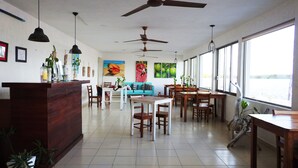 Un comedor amplio con varias mesas y sillas de madera, ventiladores de techo y ventanas grandes que ofrecen vistas al exterior.