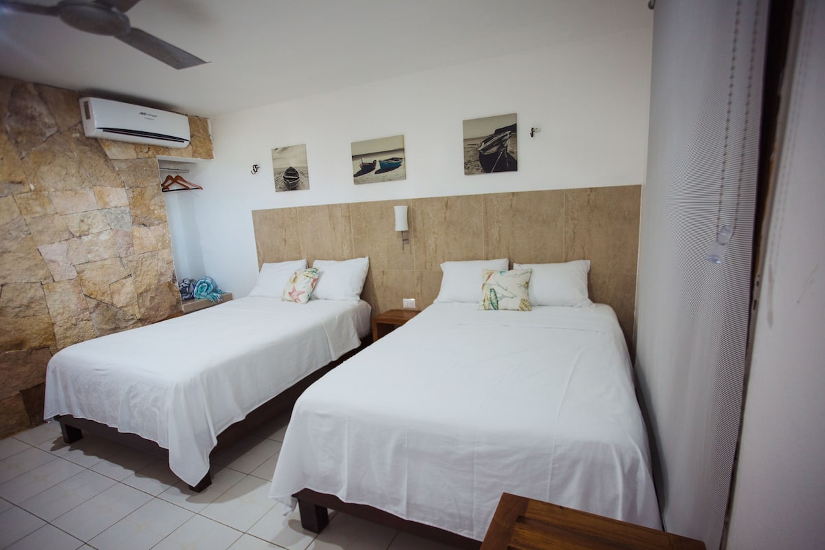 Habitación de hotel con dos camas individuales, pared de piedra, unidad de aire acondicionado y tres cuadros colgados.