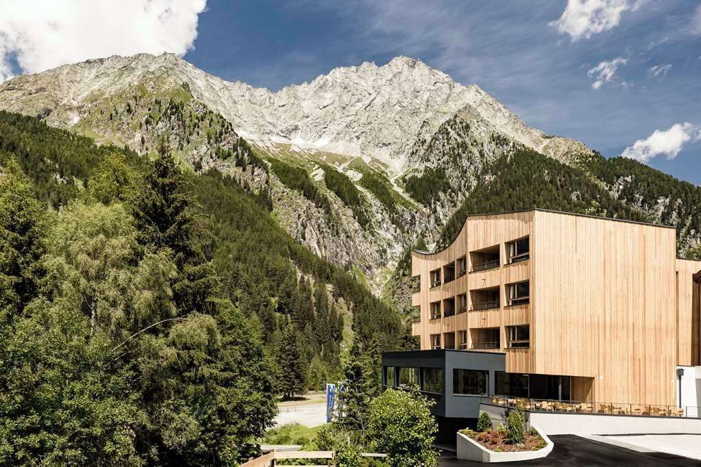 Un edificio moderno con fachada de madera, rodeado de abundante vegetación y montañas al fondo.