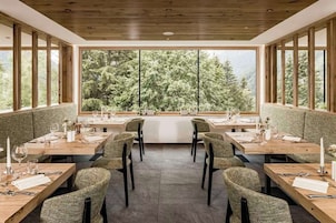Zona pranzo con tavoli e sedie in legno, ampie finestre vista alberi.