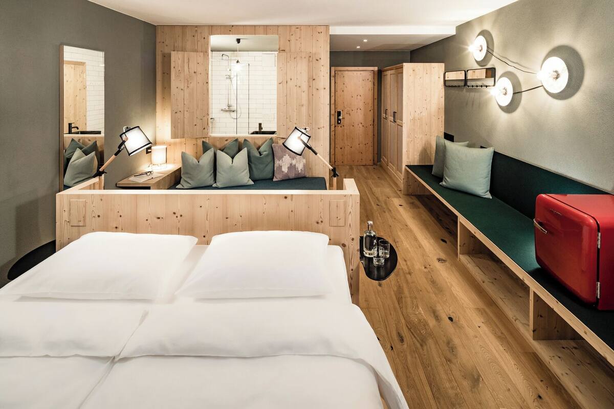 Une chambre d’hôtel moderne avec un plancher en bois, un lit recouvert de draps blancs, un coin salon avec des coussins, une petite table avec une lampe, une armoire et un luminaire fixé au mur.