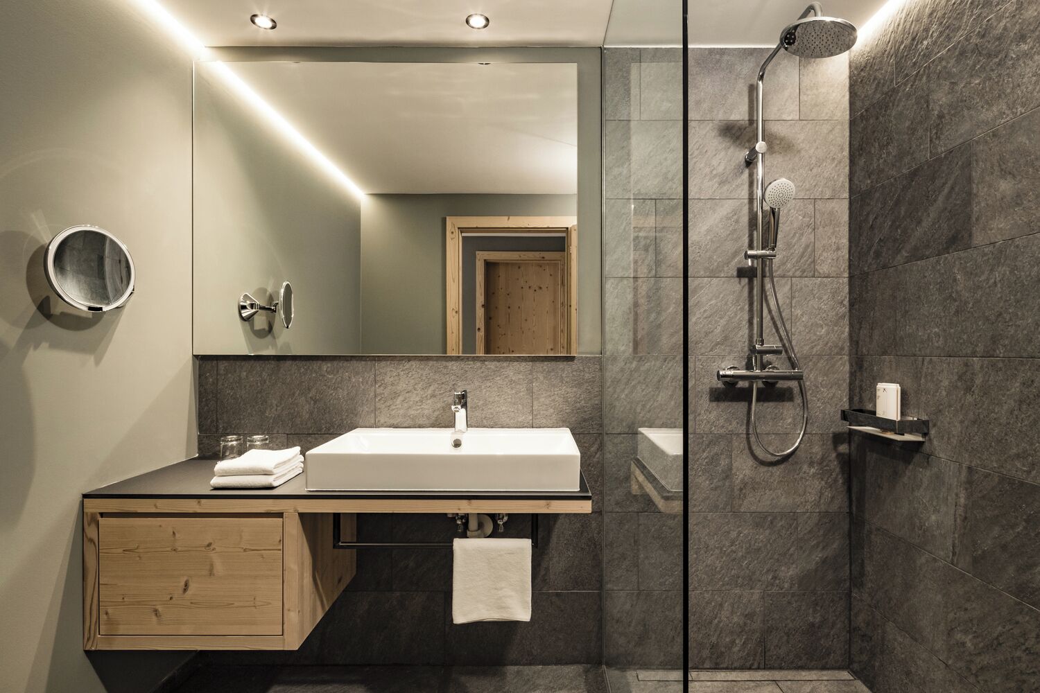 Un baño moderno con un espejo grande, un lavamanos blanco, una ducha y un mueble de tocador de madera.