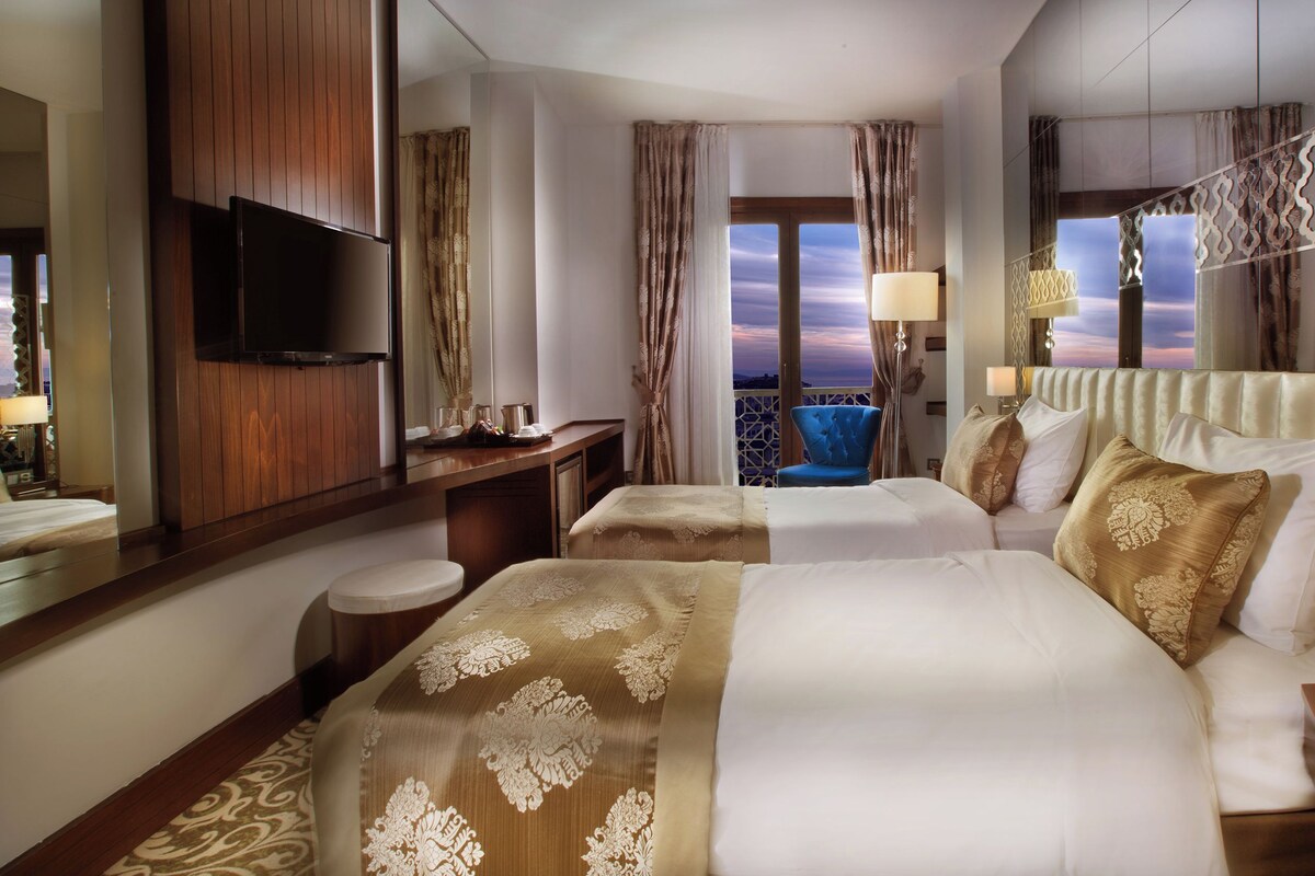 Habitación de hotel con dos camas, televisión, balcón con vistas y baranda decorativa.