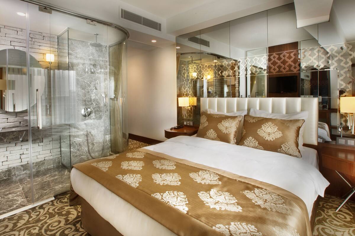 A hotel room with a glass-enclosed shower, a large bed with a patterned bedspread, a headboard with built-in lighting, and a decorative mirror with a wavy frame.
