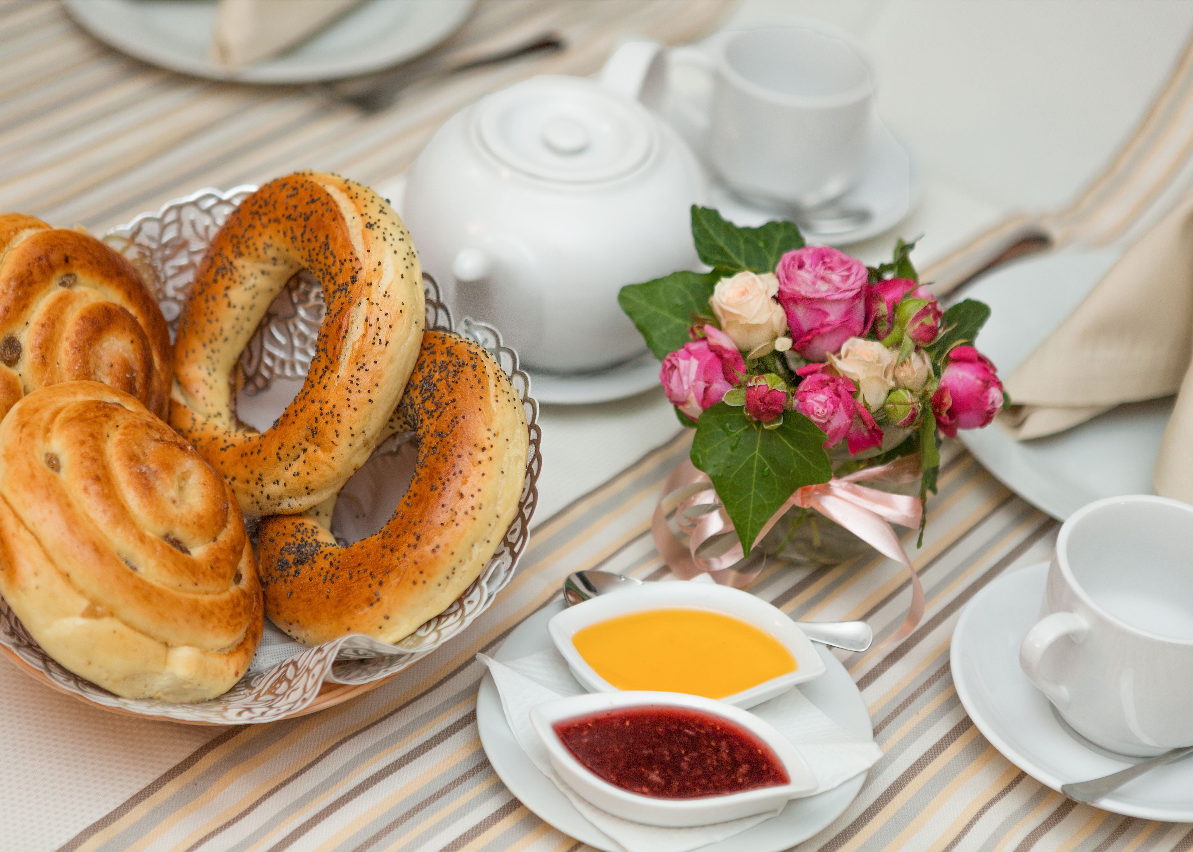 Una mesa con bagels, una tetera, un ramo de flores y dos recipientes pequeños de mermelada.