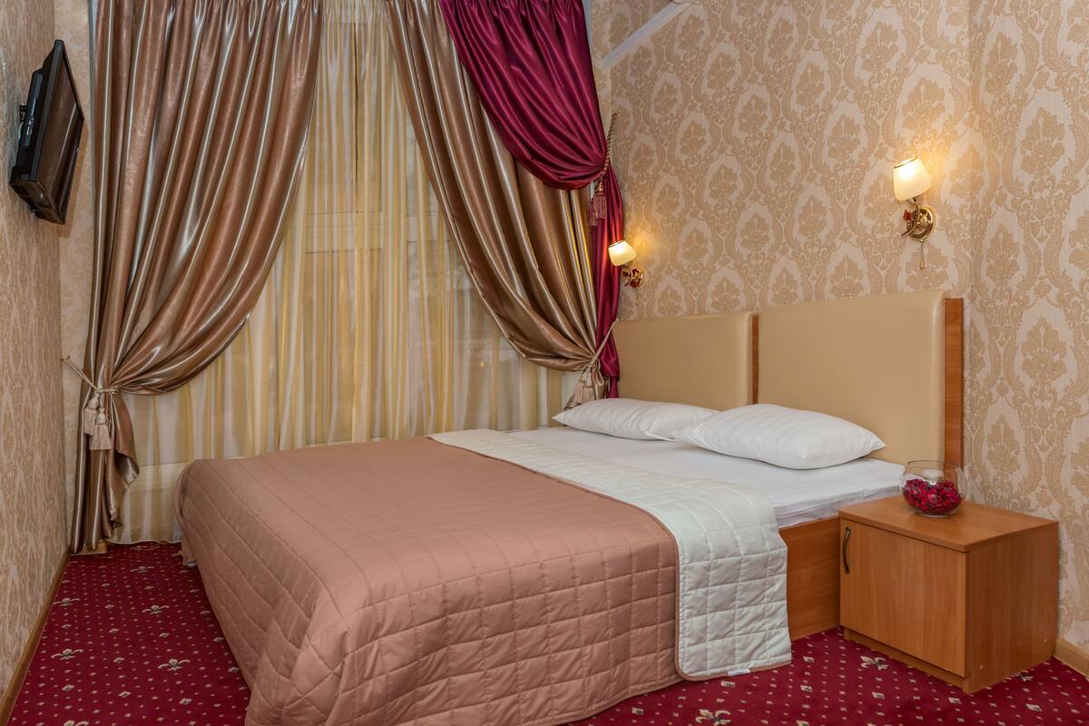 A neatly made bed with a pink quilt, a wooden nightstand with a lamp, and a wall-mounted TV.