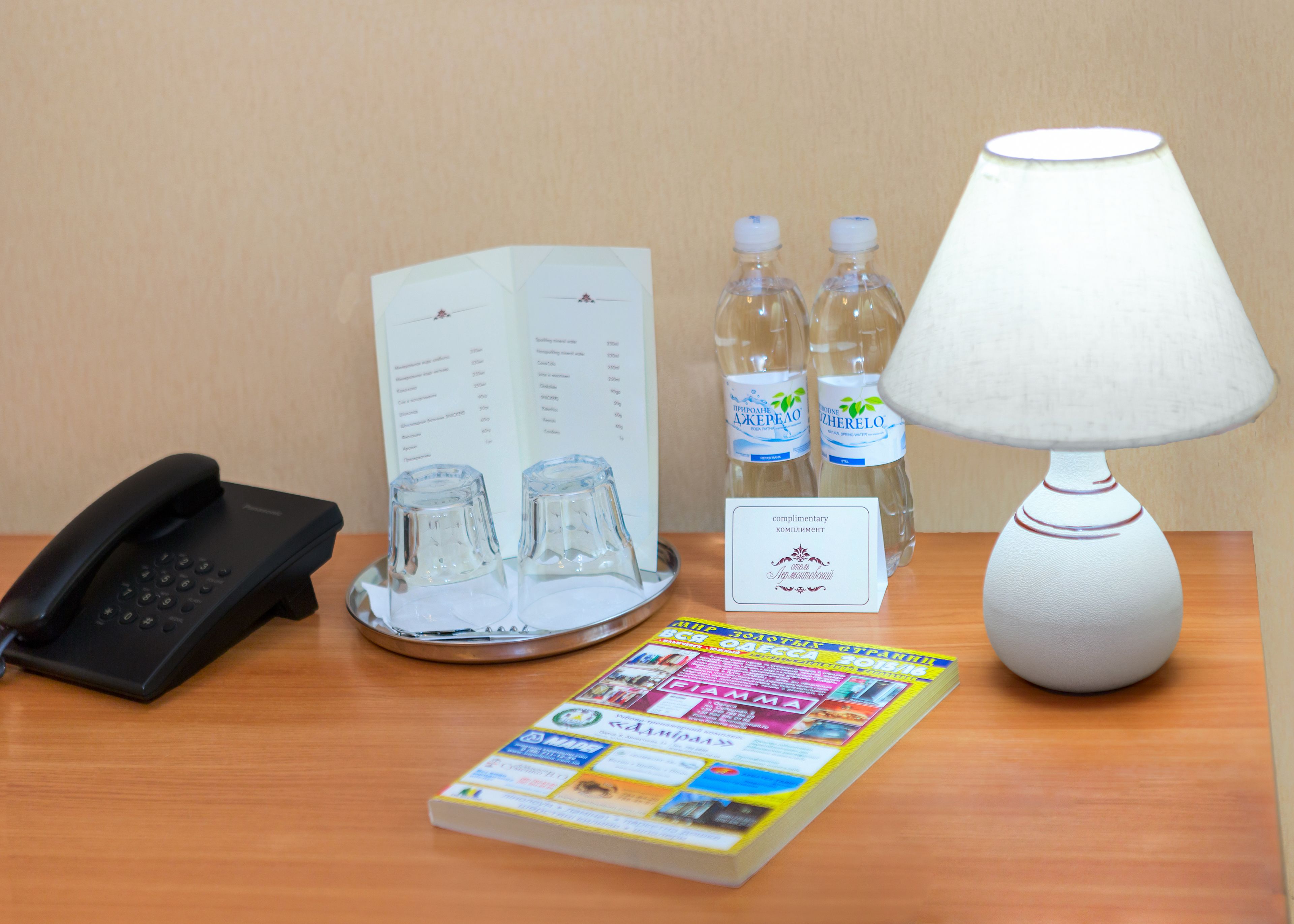 A hotel room with a black telephone, a glass water bottle, a white lamp, a menu, and a magazine on the table.