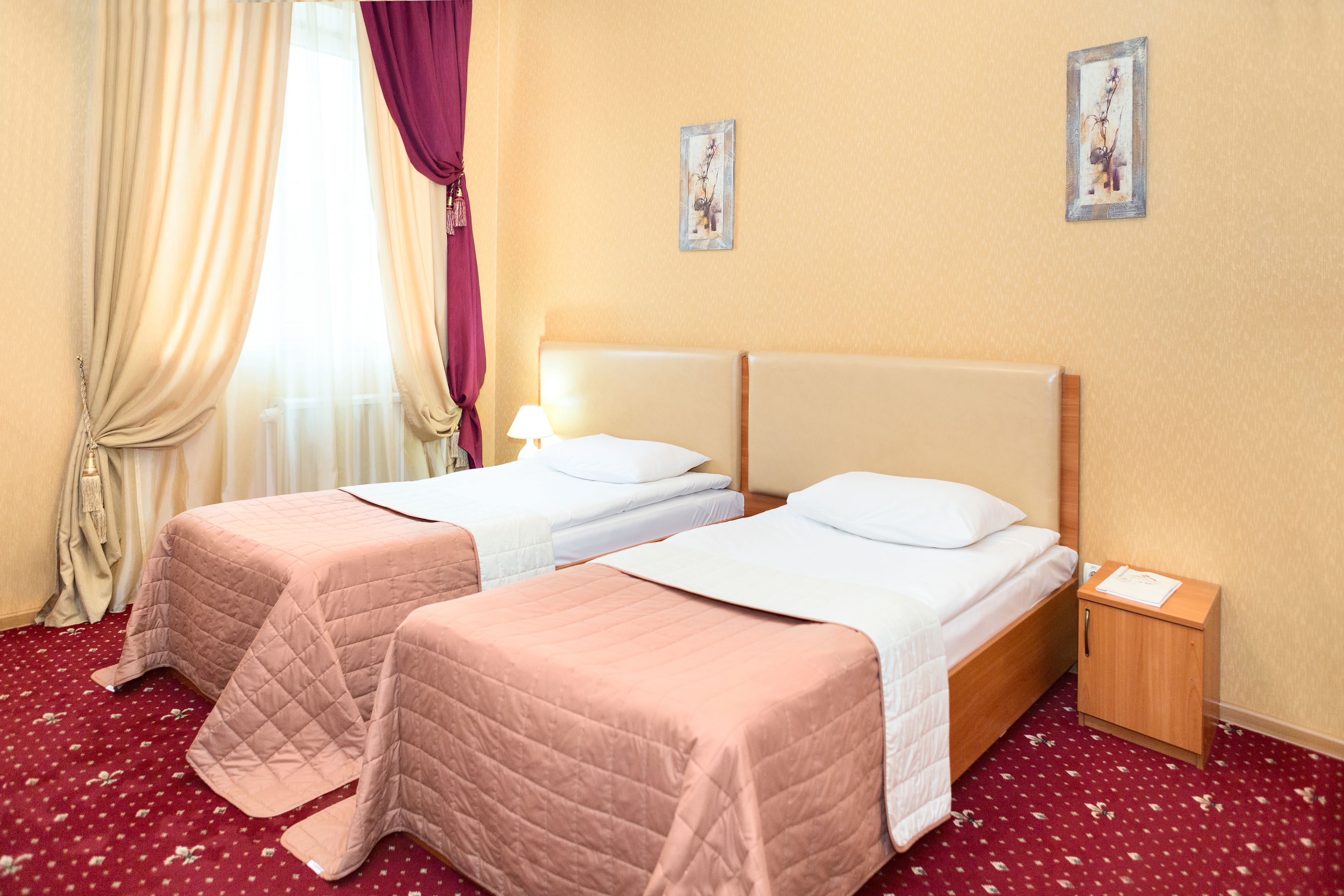 A hotel room with two beds, a wooden nightstand, a red carpet, and a window with curtains.