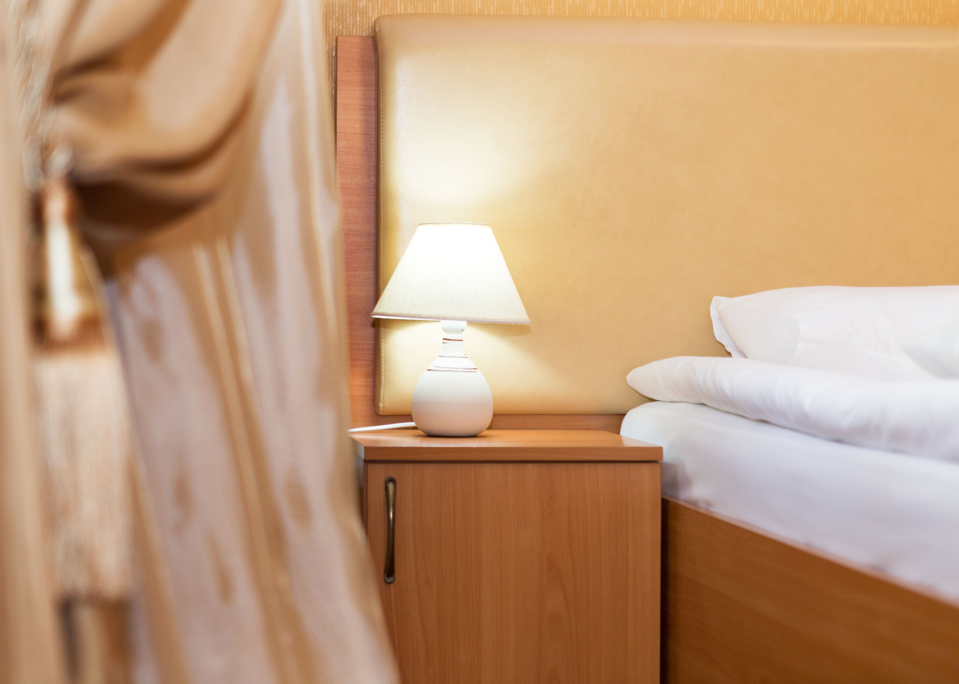 Una cama bien hecha con sábanas blancas, una mesita de noche de madera con una lámpara blanca y un cabecero de madera.