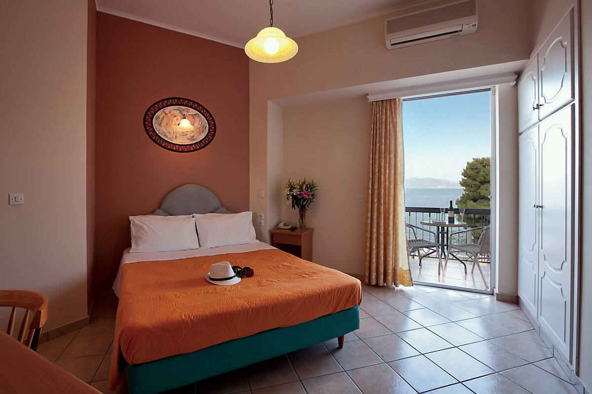 A hotel room with a bed, a small table with a vase, a balcony with a table and chairs, and a view of the sea.