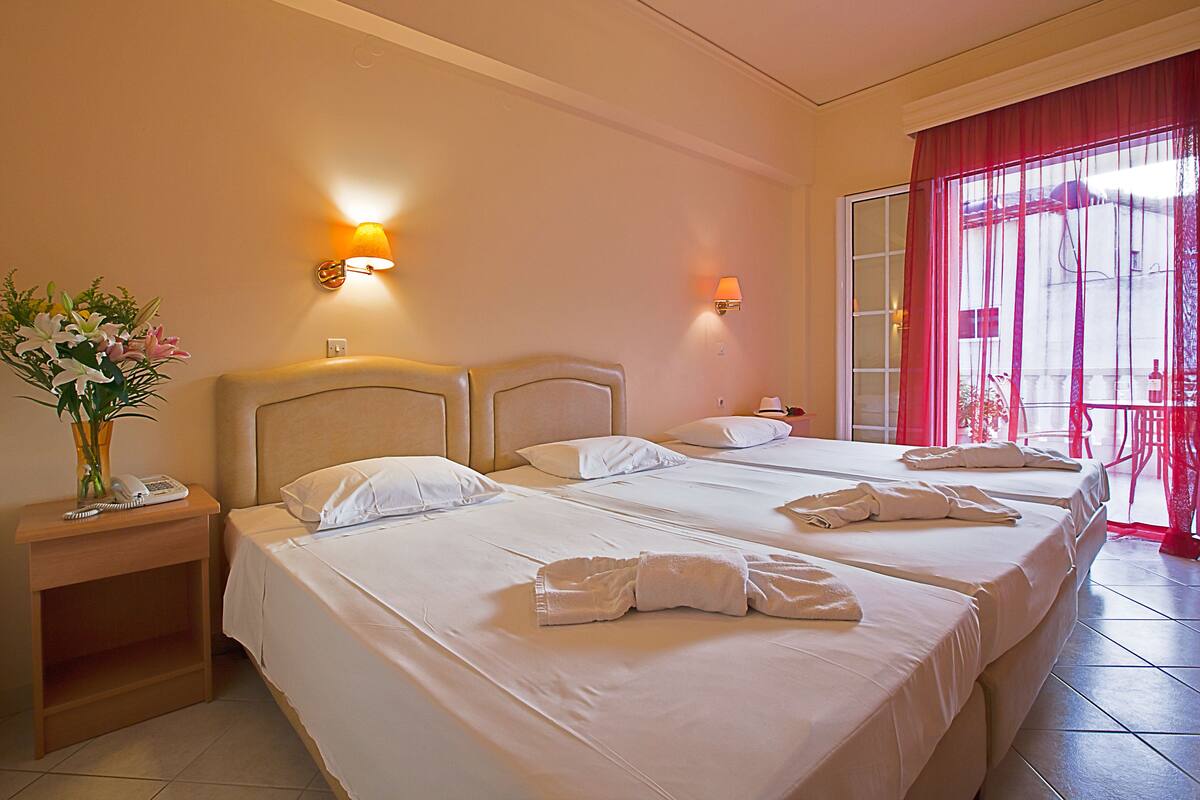 A hotel room with two beds, a nightstand, a vase with flowers, and a window with red curtains.