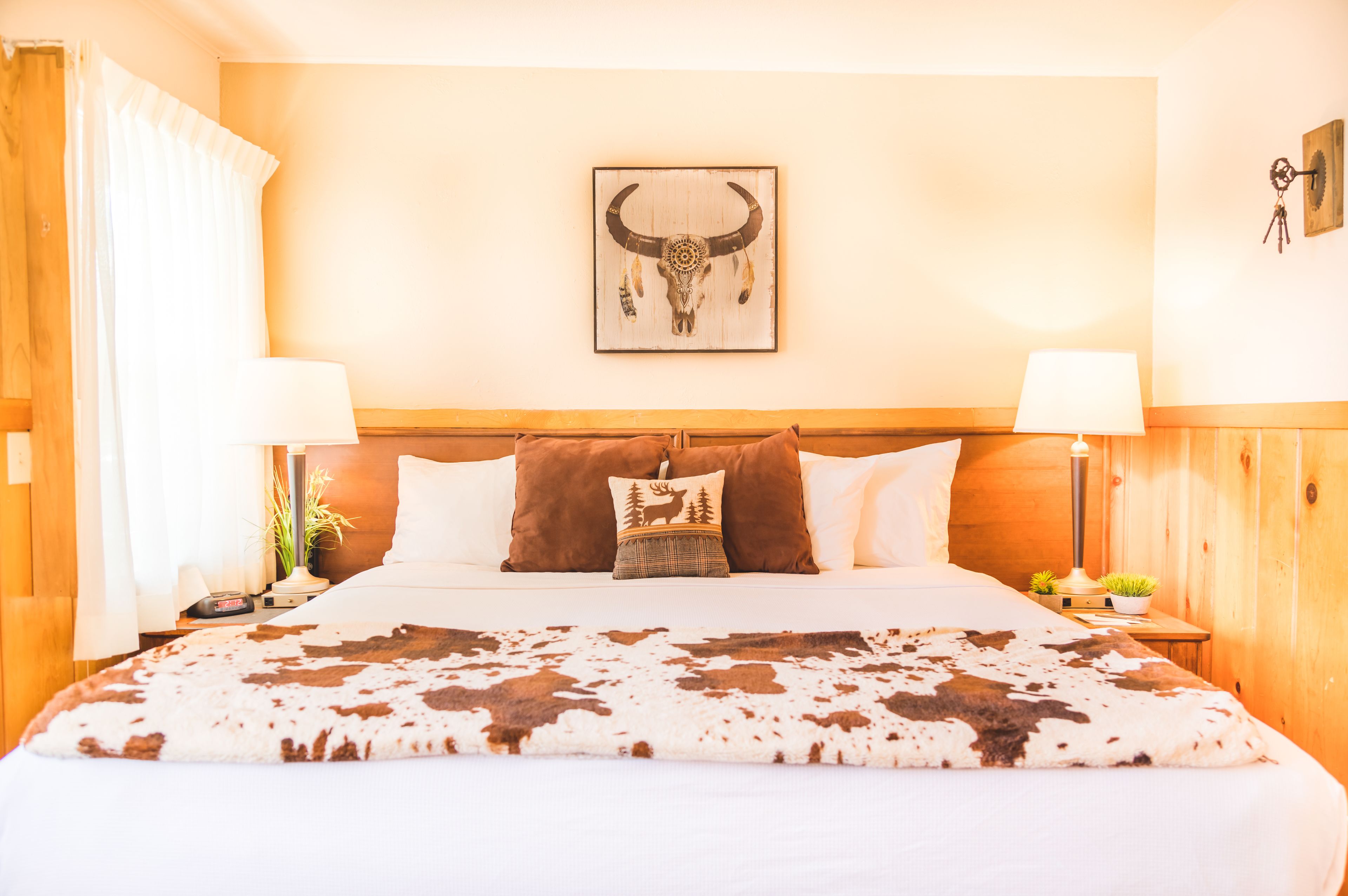 Une chambre d’hôtel avec une tête de lit en bois, un grand lit recouvert d’une couverture en peau de vache, deux lampes, une décoration murale représentant un crâne de taureau et une table de chevet agrémentée d’une plante et d’une lampe.