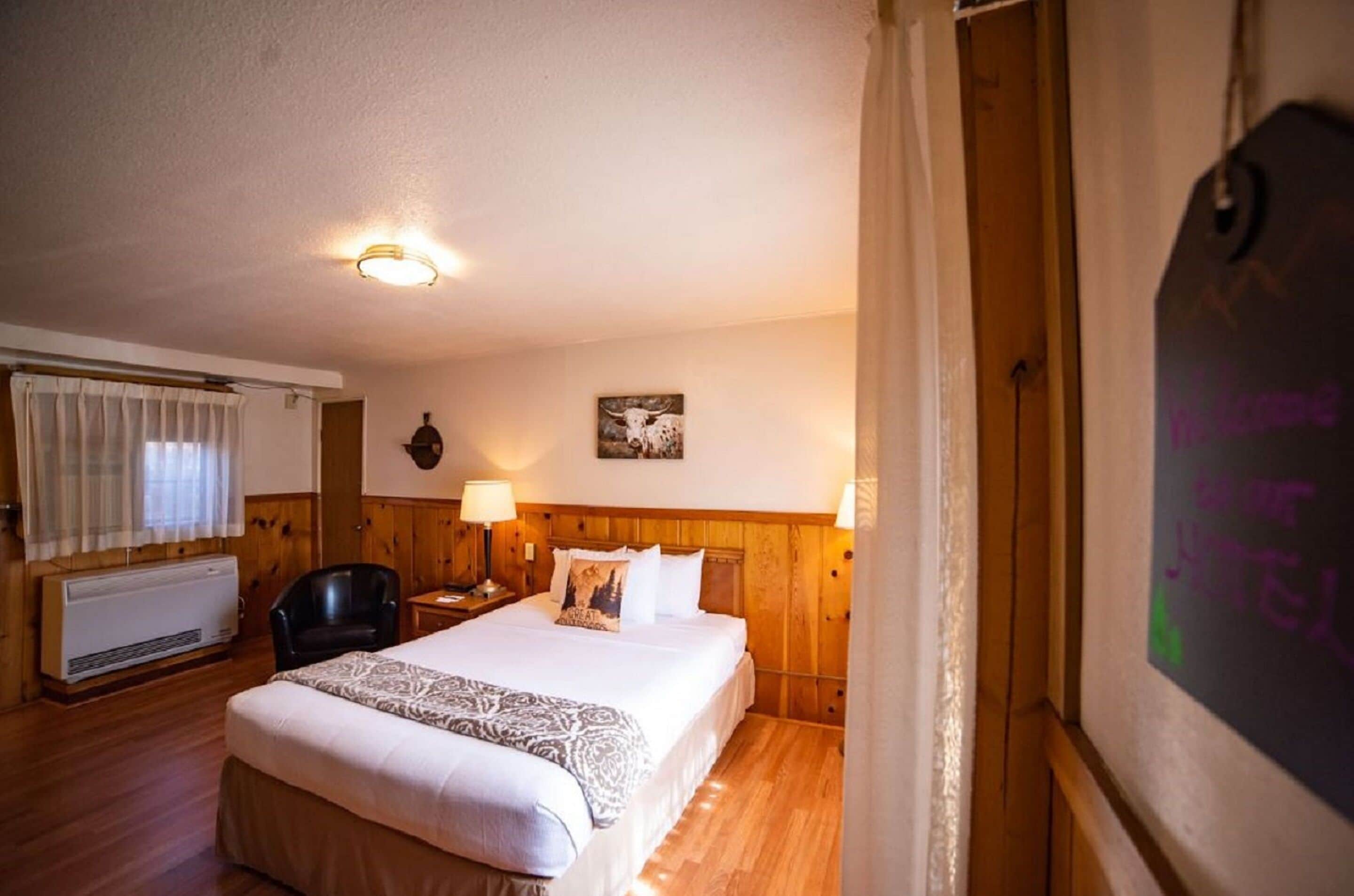 A hotel room with a wooden headboard, a bed with white linens, a nightstand with a lamp, a window with curtains, and a wall-mounted heater.