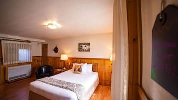 A hotel room with a wooden headboard, a bed with white linens, a nightstand with a lamp, a window with curtains, and a wall-mounted heater.
