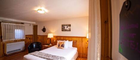 A hotel room with a wooden headboard, a bed with white linens, a nightstand with a lamp, a window with curtains, and a wall-mounted heater.