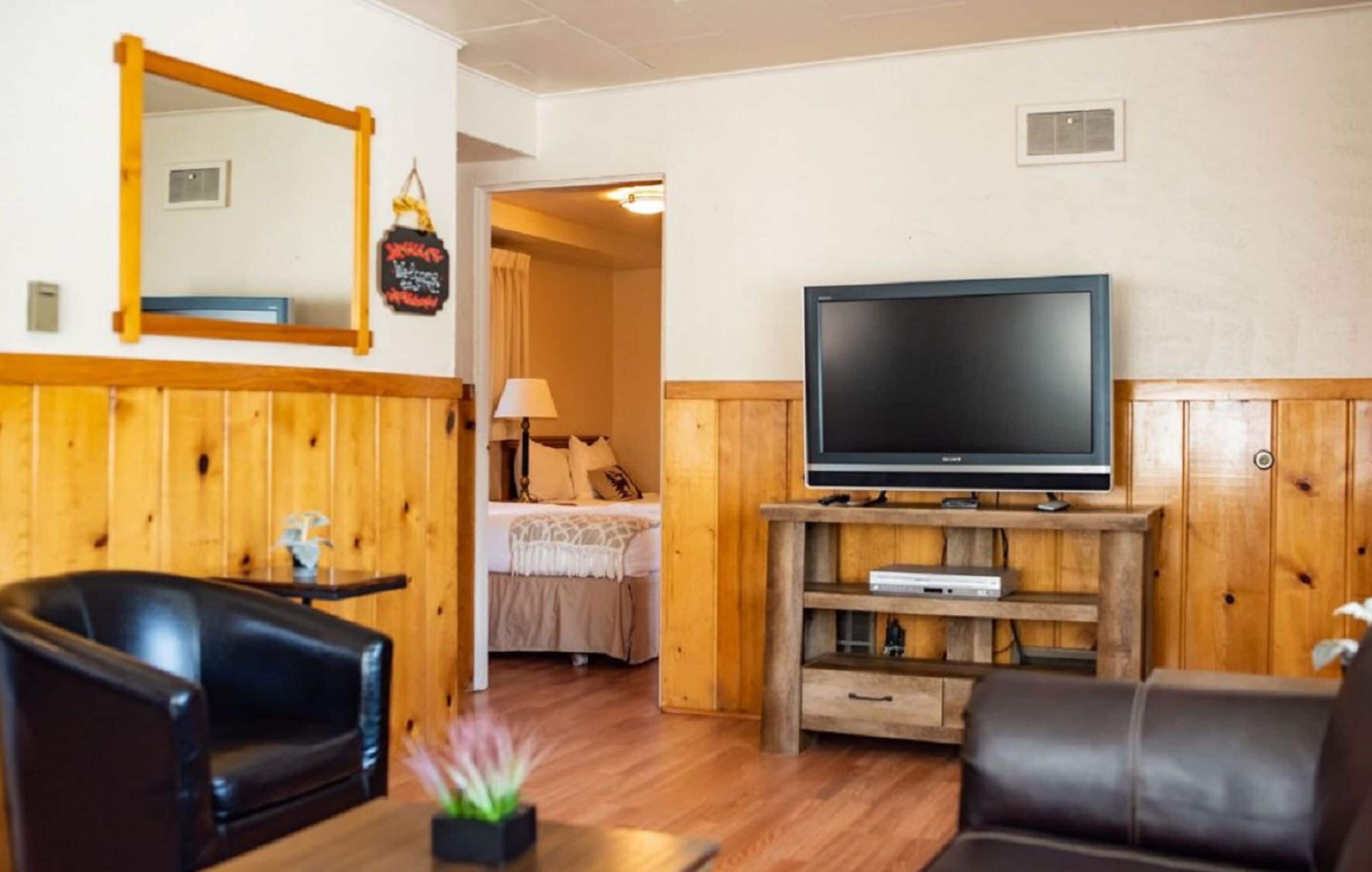 Um quarto com parede revestida de madeira, uma televisão sobre suporte de madeira, uma cama com colcha branca e uma poltrona de couro preto.