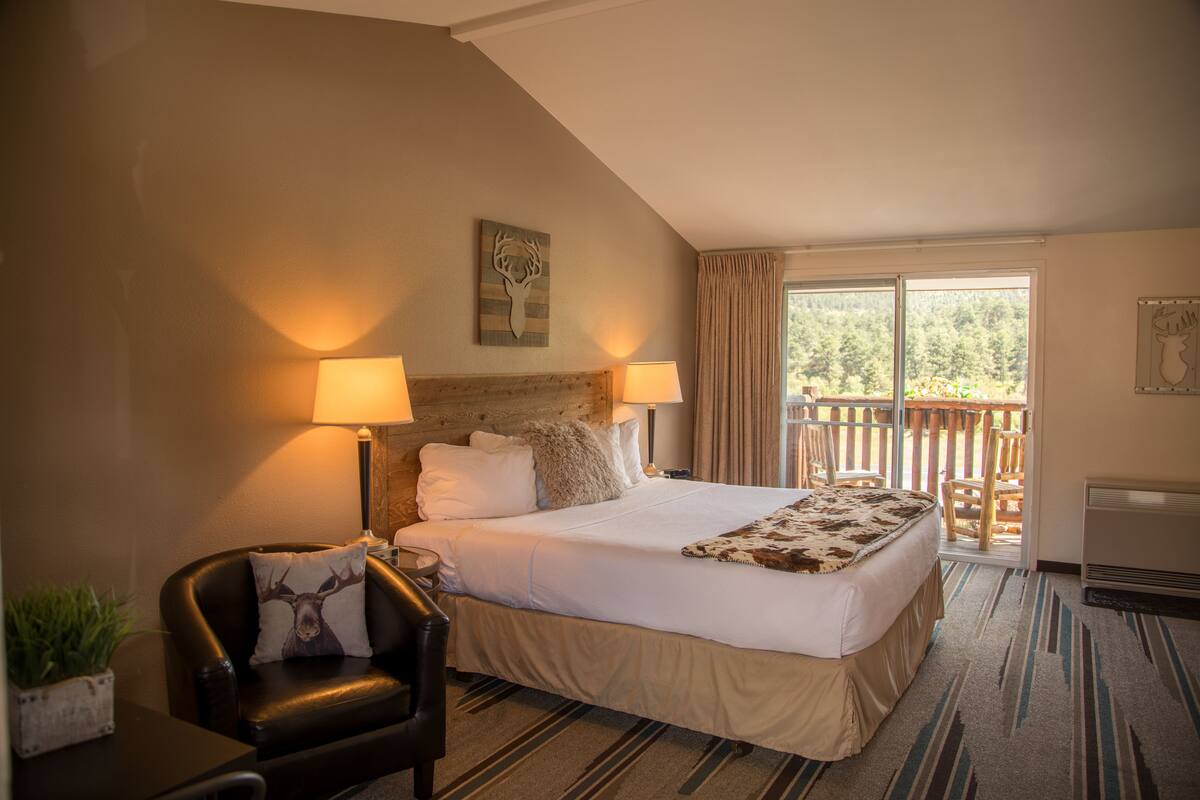 A hotel room with a large bed, two bedside lamps, a wooden headboard, a balcony with a view of trees, and a small table with a plant.