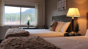 A hotel room with two beds, a nightstand with a lamp, a window with curtains, a small table with a plant, and a wall-mounted artwork. - Alpine Trail Ridge Inn (Estes Park)