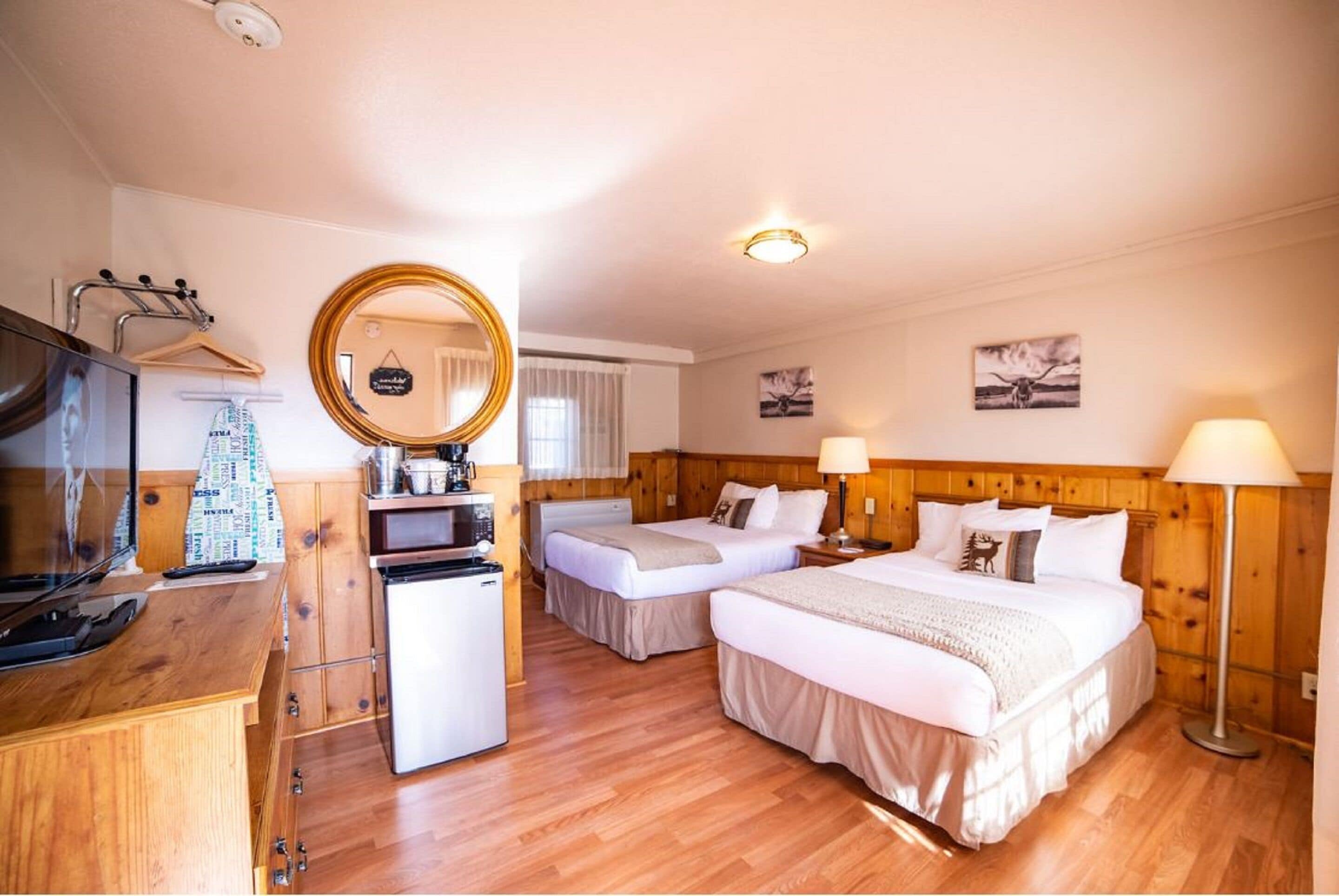 Une chambre d’hôtel avec deux lits, une tête de lit en bois, une petite kitchenette, une télévision, un miroir et une fenêtre avec des rideaux.