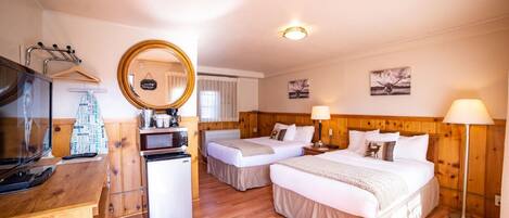 Une chambre d’hôtel avec deux lits, une tête de lit en bois, une petite kitchenette, une télévision, un miroir et une fenêtre avec des rideaux.