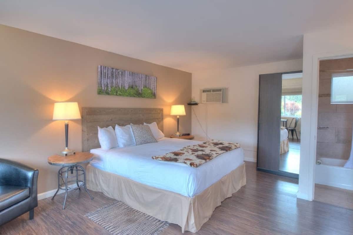 A bedroom with a bed, two bedside lamps, a chair, a painting on the wall, and a bathroom visible through an open door.