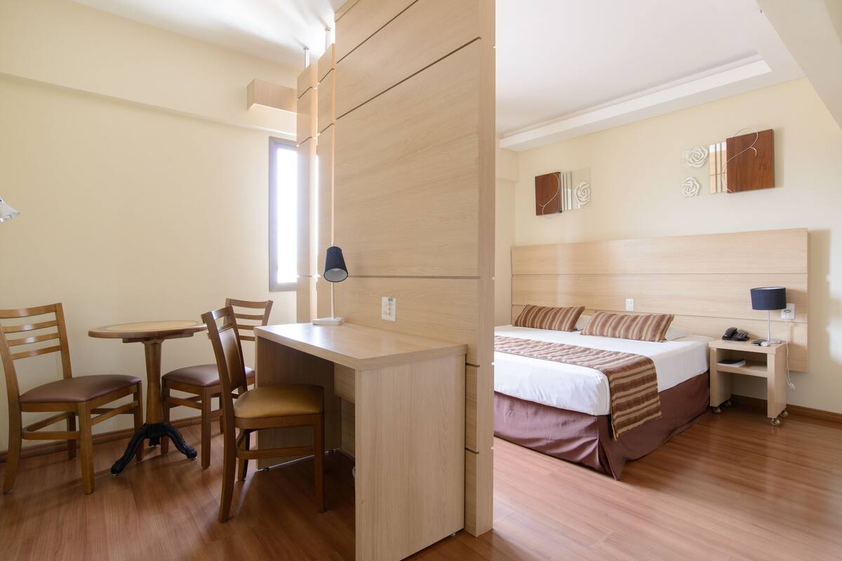 A hotel room with a wooden desk, chairs, a bed with a headboard, bedside tables, and wall art.