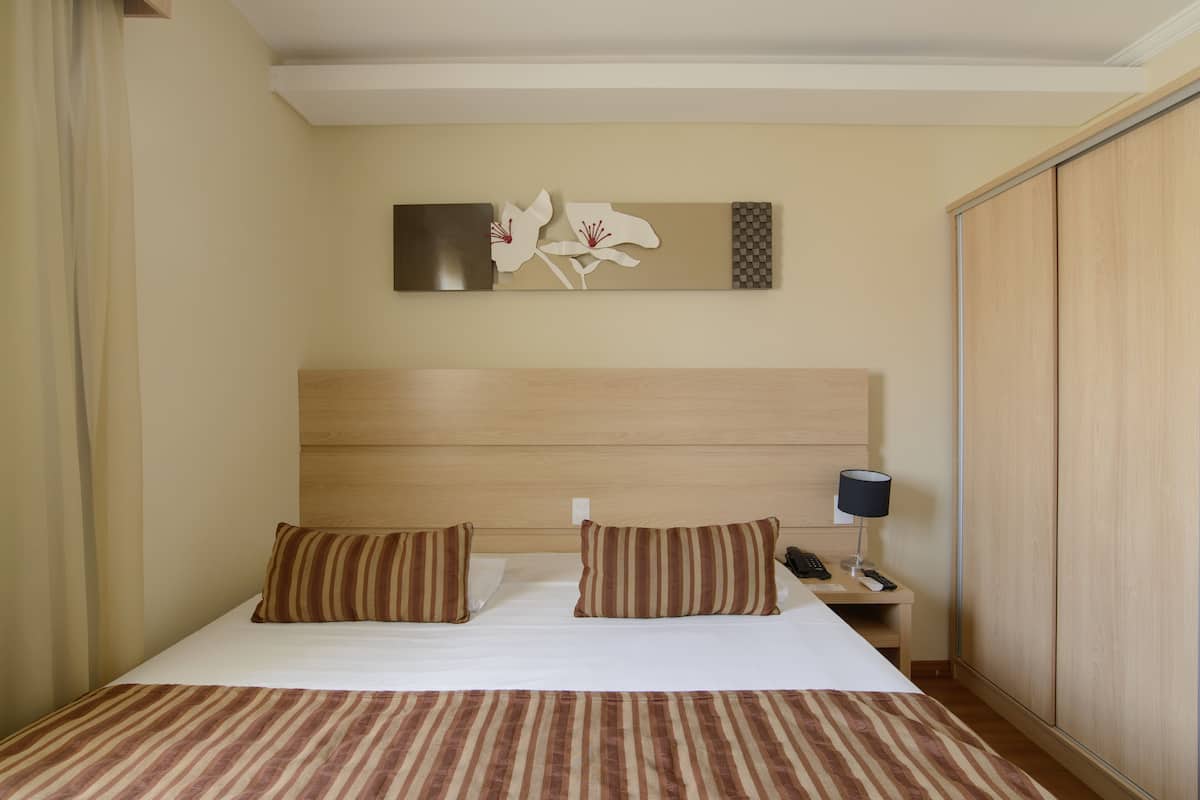 A hotel room with a bed, two pillows, a nightstand with a lamp, a phone, and a wall-mounted artwork.