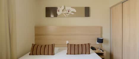 A hotel room with a bed, two pillows, a nightstand with a lamp, a phone, and a wall-mounted artwork.