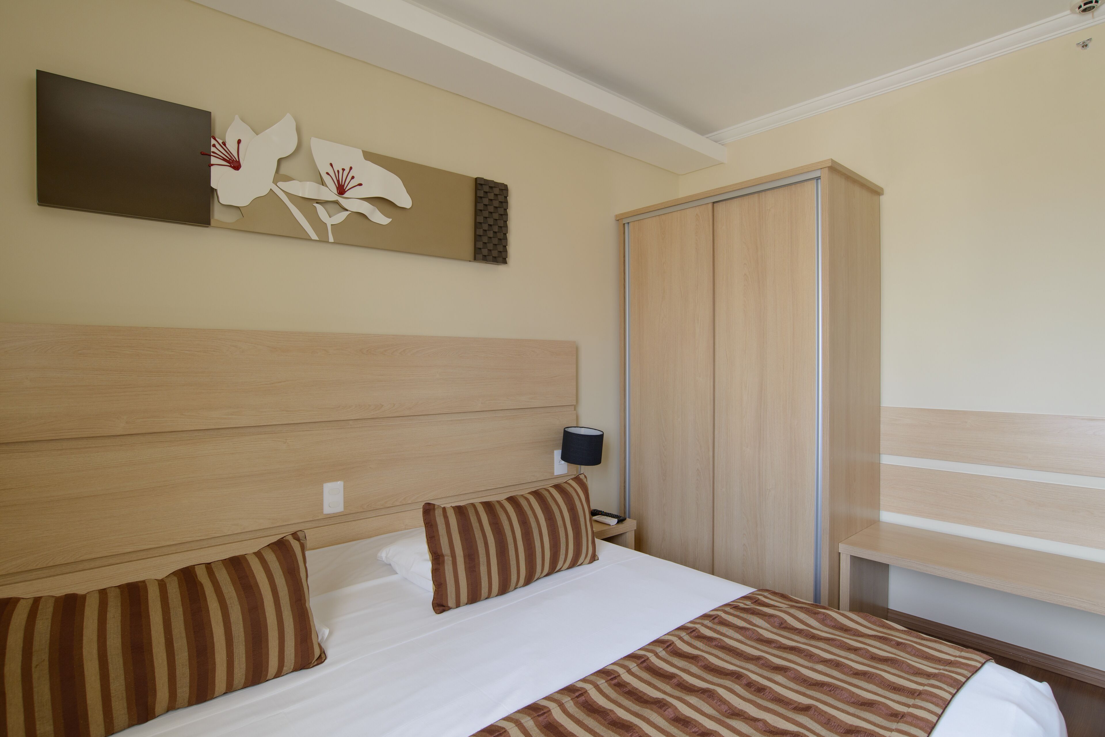 Habitación de hotel con una cama grande, cabecera de madera, mesita de noche con lámpara, armario y una obra de arte colgada en la pared.