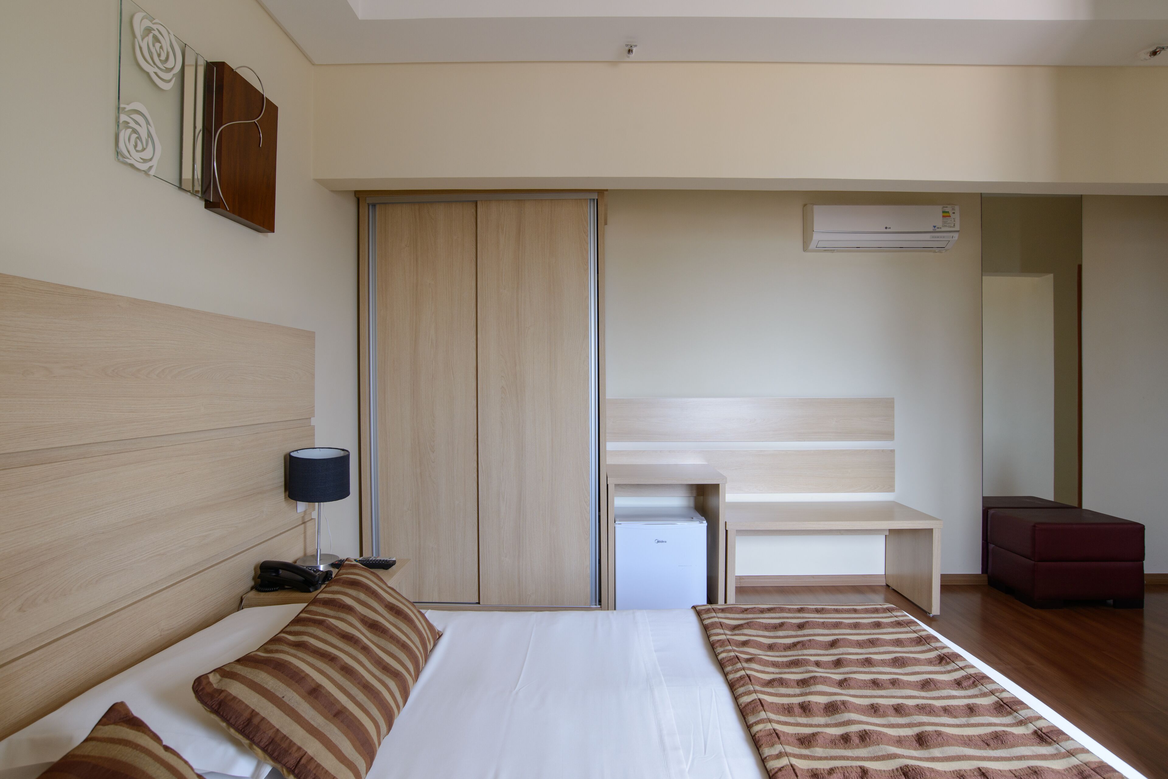 A hotel room with a bed, bedside table, lamp, wardrobe, and a small desk with a chair.