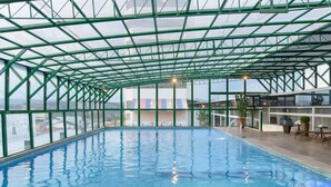 An indoor swimming pool with a glass ceiling and a view of the cityscape. - Faro Hotel (Atibaia)
