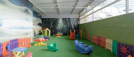 An indoor play area with artificial grass, colorful play structures, and a mural on the wall.