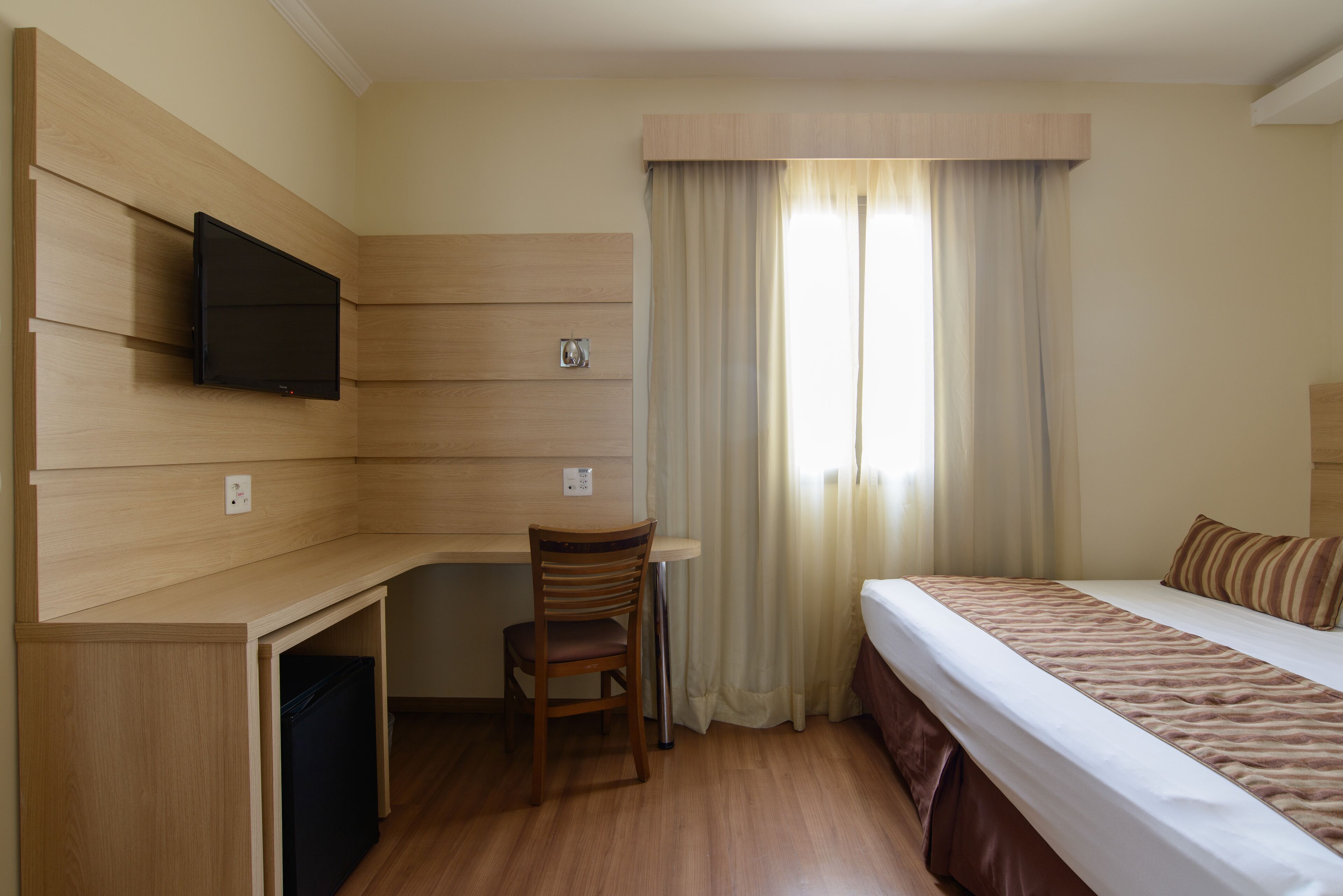 Habitación de hotel con cama, escritorio con silla, televisor montado en la pared y ventana con cortinas.