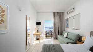 A hotel room with a bed, a sofa, a desk with chairs, a TV, and a balcony with a view of the sea. - Hotel Flisvos (Sitia)